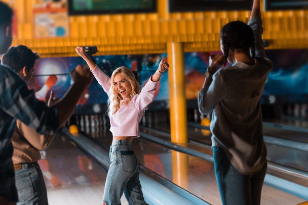 vzrušené blondýny dívka ukazuje vítězný gesto při stání v blízkosti bowling uličky s multikulturními přáteli - Fotografie, Obrázek