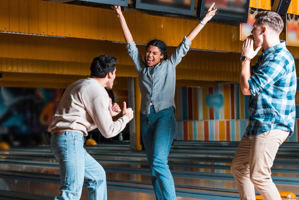 glückliches afrikanisch-amerikanisches Mädchen, das mit den Händen in der Luft neben multikulturellen Freunden steht, die Daumen nach oben zeigen und im Bowlingclub schreien - Foto, Bild