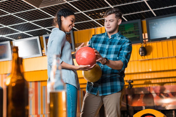 selectieve focus van knappe man tonen bowling bal naar lachende Afrikaans Amerikaans meisje  - Foto, afbeelding