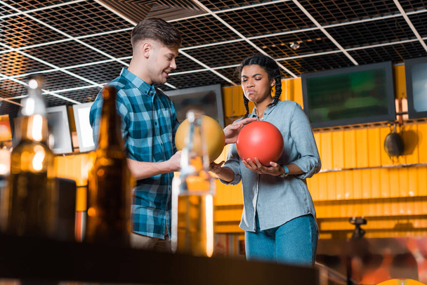 selective focus of smiling man talking to displeased african american girl while holding bowling balls - Foto, afbeelding