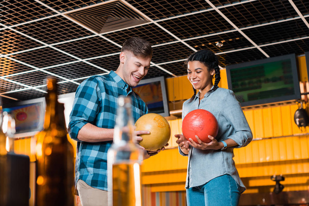 selective focus of interracial couple talking and smiling while holding bowling balls - Foto, afbeelding