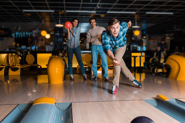 sonriente joven lanzando bowling bowl en skittle alley cerca de amigos multiculturales
 - Foto, Imagen
