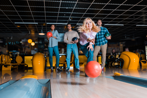 smiling blonde girl throwing bowling ball on skittle alley near multicultural friends - Photo, Image