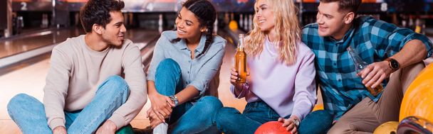 tiro panorâmico de amigos multiculturais felizes com garrafas de cerveja sentado e falando no beco skittle no clube de boliche
 - Foto, Imagem