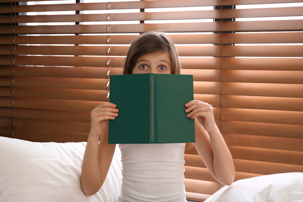 Emotional preteen girl with book on bed near window - Фото, зображення
