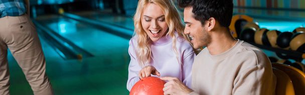 panoramic shot of cheerful couple looking at bowling ball while sitting in bowling club - Photo, Image