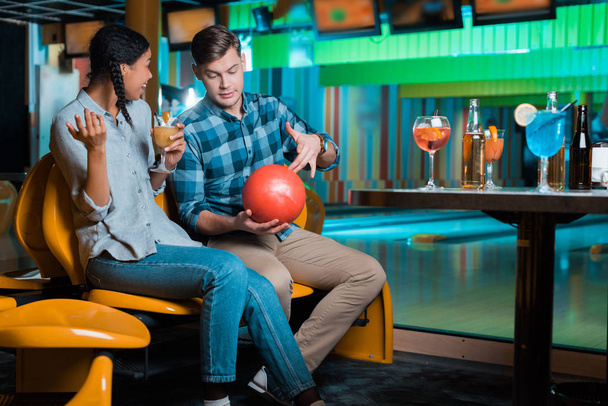 young man showing bowling ball to smiling african american girl holding cocktail - Photo, Image