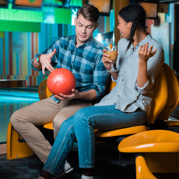usmívající se africká americká dívka drží sklenici koktejlu, zatímco přítel ukazuje bowling koule - Fotografie, Obrázek