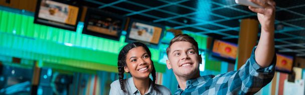 plan panoramique de couple interracial heureux prenant selfie sur smartphone tout en étant assis dans le club de bowling
 - Photo, image
