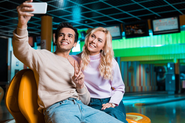 szczęśliwy młody człowiek pokazując gest zwycięstwa siedząc w bowilng klub i selfie z uśmiechniętej dziewczyny - Zdjęcie, obraz