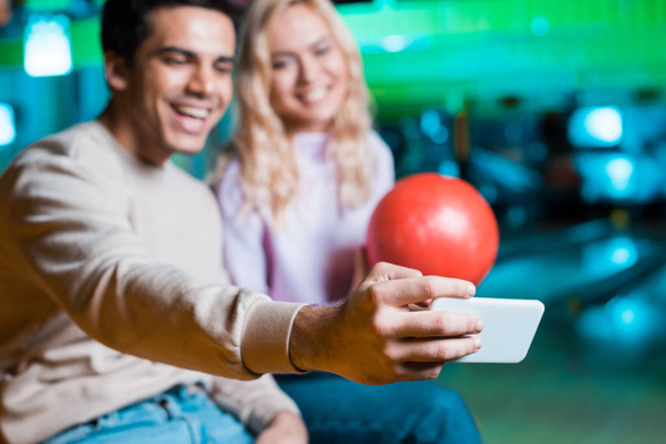 selectieve focus van glimlachend meisje houden bowling bal, terwijl vrolijke vriend het nemen van selfie op de smartphone - Foto, afbeelding
