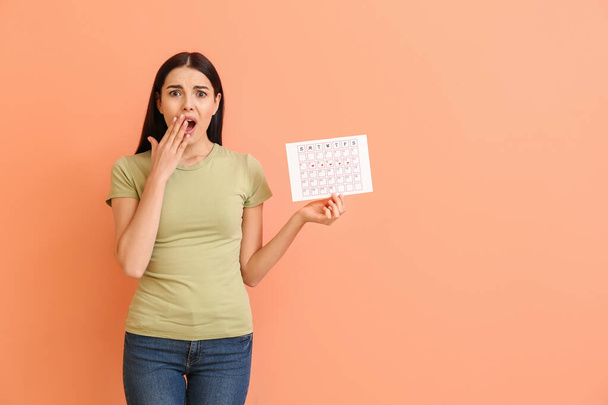 Schockierte Frau mit Menstruationskalender auf farbigem Hintergrund - Foto, Bild