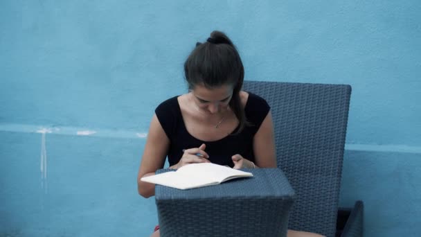 Young woman sits on sunbed, makes notes in diary, uses mobile, blue background - Záběry, video