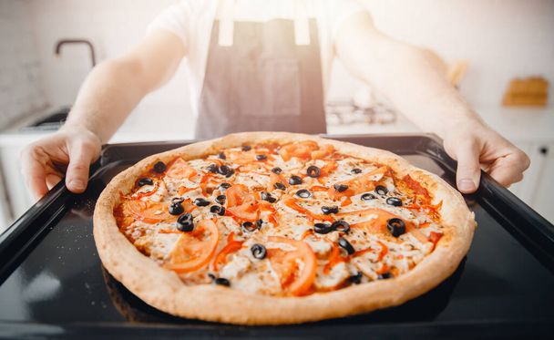 Pizza italiana fresca caliente en bandeja para hornear del horno, chef en delantal sostiene en las manos, la luz del sol
 - Foto, imagen