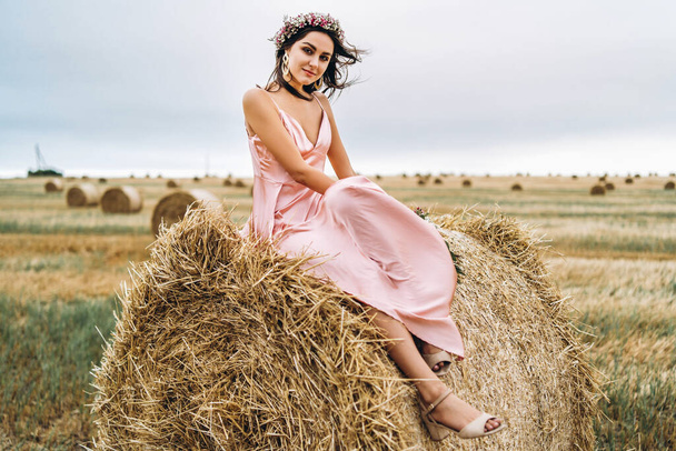 Young brunette in a pink satin dress sitting on a hay bale. A wo - Foto, Imagen