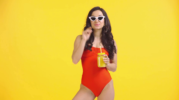alegre chica en traje de baño beber jugo y agitar la mano aislado en amarillo
 - Metraje, vídeo
