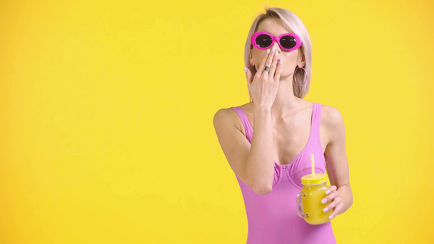 χαρούμενο κορίτσι κουνώντας το χέρι, φυσώντας φιλί αέρα, πίνοντας χυμό απομονωμένο στο κίτρινο - Πλάνα, βίντεο
