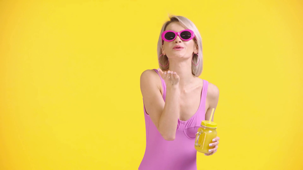 Ευτυχισμένο κορίτσι κουνώντας το χέρι, φυσώντας αέρα φιλί και πόσιμο χυμό που απομονώνονται σε κίτρινο - Πλάνα, βίντεο