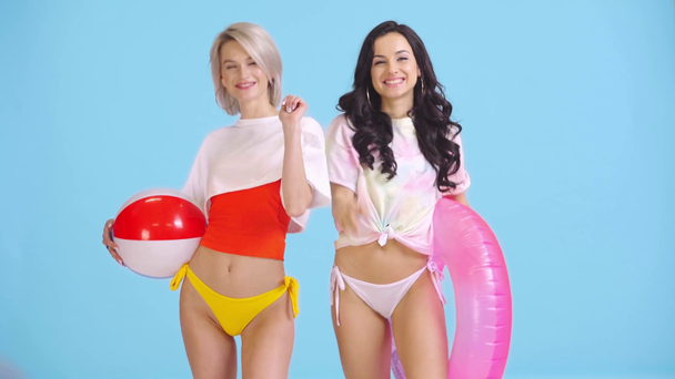 smiling girls with inflatable ball and swim ring waving hands isolated on blue - Footage, Video