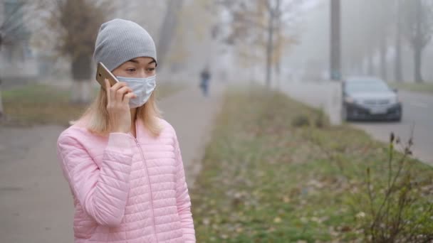 Protezione contro il Coronavirus. Una giovane donna in una maschera di protezione medica sta con uno smartphone in una strada della città in Europa. Sintomi di coronavirus si sono presentati. - Filmati, video