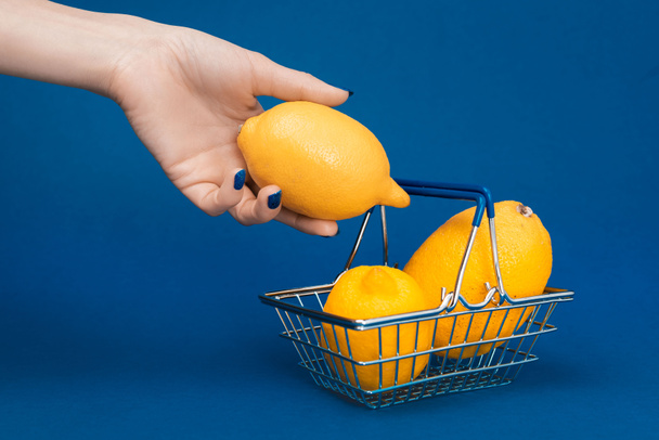 cropped view of woman putting lemon in shopping basket on blue background  - 写真・画像