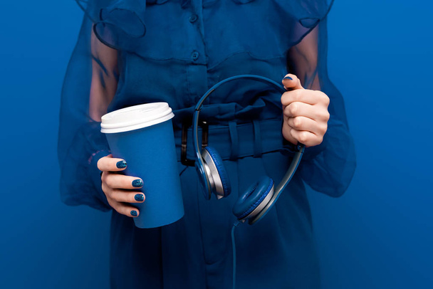 vista cortada de mulher segurando copo de papel e fones de ouvido no fundo azul
  - Foto, Imagem