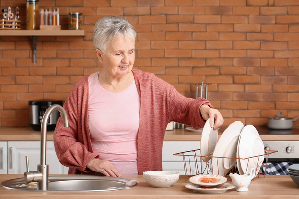 Starsza kobieta zmywa naczynia w kuchni - Zdjęcie, obraz