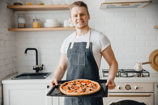 Chef masculino europeo sonríe y sostiene bandeja para hornear con pizza italiana caliente en cocina luminosa
 - Foto, imagen