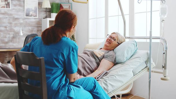En casa de retiro brillante anciana acostada en la cama del hospital habla con una cuidadora
 - Foto, Imagen