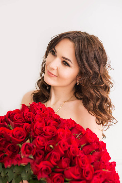 Όμορφη νεαρή γυναίκα με μπουκέτο κόκκινα τριαντάφυλλα που απομονώνονται σε λευκό φόντο. Μακρύ κυματιστό χτένισμα. Πορτρέτο στούντιο - Φωτογραφία, εικόνα