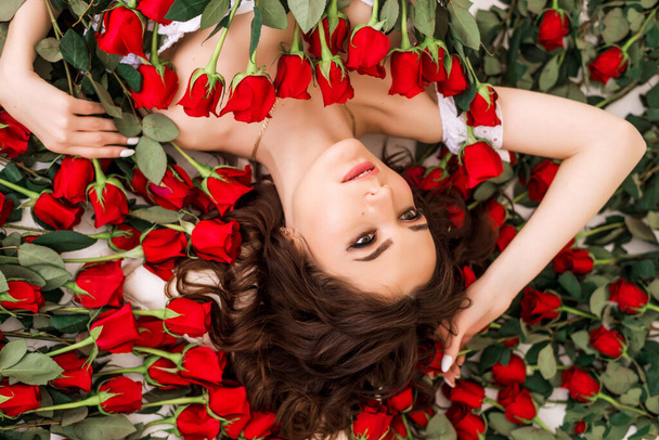 Güller içinde güzel esmer bir kadının yakın plan portresi. Kız kırmızı güllerle çevrili yatıyor. Profesyonel makyaj malzemesi. Güzellik salonu ve parfüm konsepti - Fotoğraf, Görsel