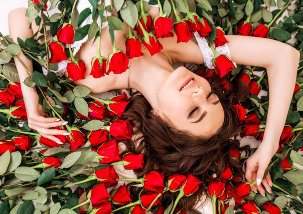 Egy gyönyörű lány portréja vörös virágokkal és rózsákkal. Szépség high fashion modell nő arc közelkép. Tökéletes bőr. Profi smink. Alakzat. Rúzs, Alapítvány, szempillaspirál reklám. - Fotó, kép