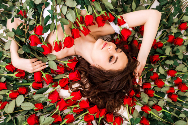Close-up portrait of a beautiful brunette woman in roses. The girl lies surrounded by red roses. Professional make-up. The concept of a beauty salon and perfume - Fotó, kép