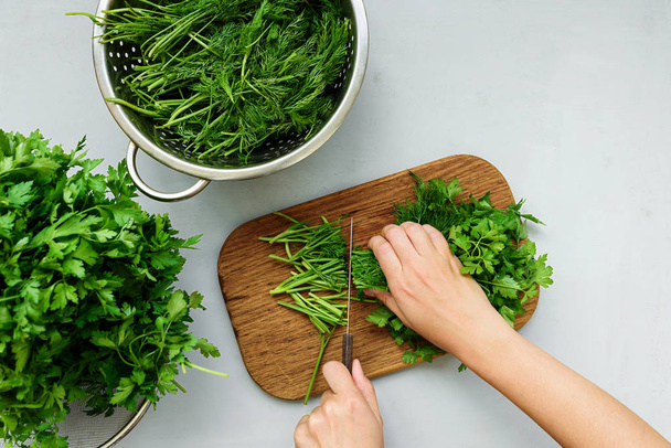 Female hands chopping fresh green parsley and dill or fennel on cutting boar on gray wooden table. Top view. Copy space. Harvesting concept - Foto, afbeelding