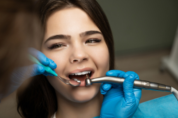 dentist in blue gloves filling the beautiful smiling woman patient's root canal using mirror under the medical lamp in clinic close up, healthcare concept - Φωτογραφία, εικόνα