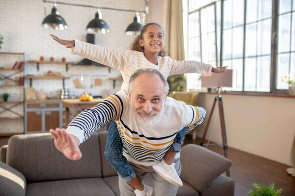 Bearded grey-haired man having fun with his dark-skinned granddaughter - Photo, image