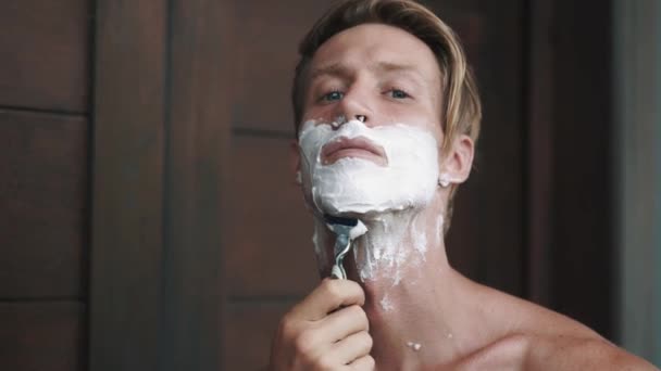Portrait of man looking at camera and shaving his beard with manual razor - Metraje, vídeo