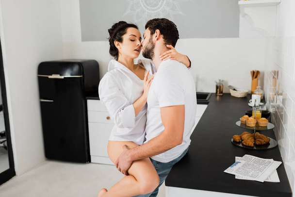 Side view of sensual woman in shirt kissing boyfriend in kitchen - Fotó, kép