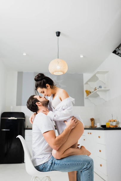 Side view of sexy girl in bra and shirt kissing boyfriend on chair in kitchen - Fotó, kép