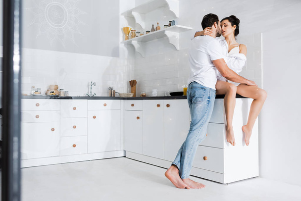Selective focus of man kissing beautiful girlfriend in shirt and bra on kitchen worktop - Фото, изображение