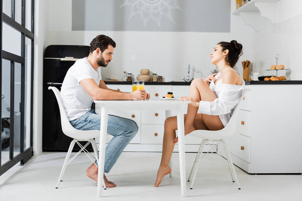 Widok z boku zmysłowa kobieta w biustonosz i koszula flirtuje z chłopakiem podczas śniadania w kuchni - Zdjęcie, obraz