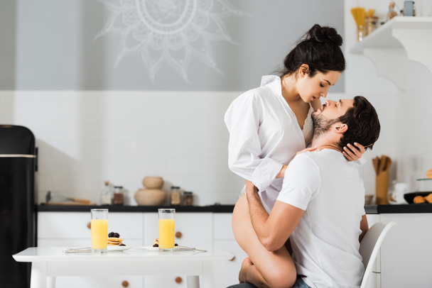 Vista lateral del hombre besándose y tocando sensual chica en lencería y camisa durante el desayuno en la cocina
 - Foto, Imagen