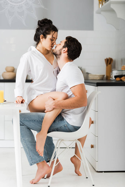 Side view of attractive woman kissing handsome boyfriend on chair in kitchen - Zdjęcie, obraz