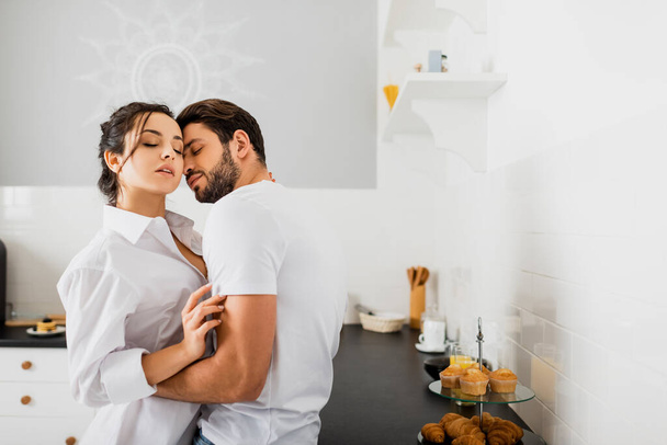 Side view of handsome man embracing sexy girlfriend in shirt near kitchen worktop - Fotografie, Obrázek