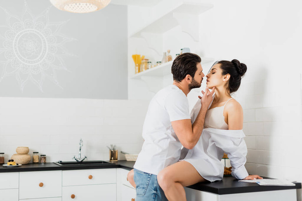 Widok z boku przystojny mężczyzna całuje sexy dziewczyna w biustonosz i koszula na stole kuchennym - Zdjęcie, obraz