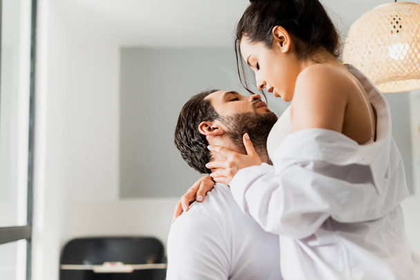 Side view of sensual woman in bra and shirt kissing handsome boyfriend in kitchen  - Fotografie, Obrázek