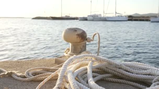Háttérvilágítás előtér napkeltekor vagy naplementekor egy kikötõbollár kötéllel és pihenõkötéllel a földön és különbözõ csónakokkal a háttérben hátsó fényben - Felvétel, videó