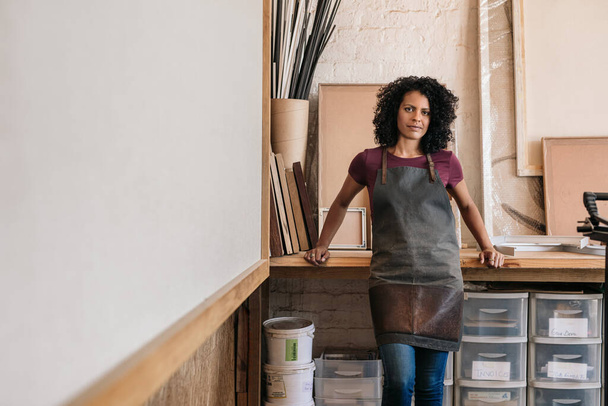 Portrait of a confident young woman wearing an apron leaning against a workbench in her picture framing studio - Φωτογραφία, εικόνα