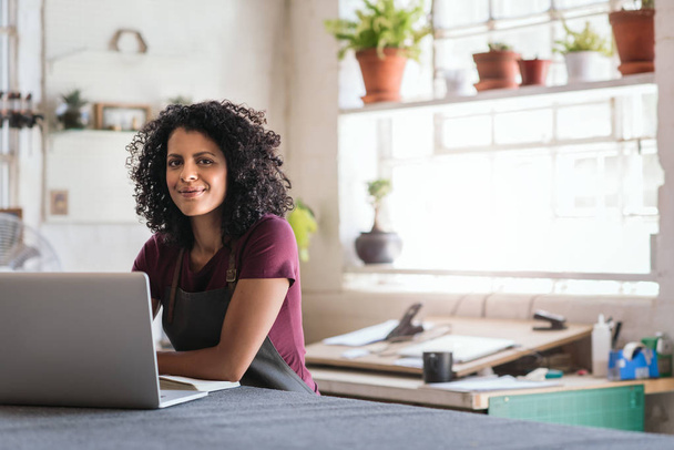 Портрет улыбающейся молодой женщины, работающей на ноутбуке, сидящей на верстаке в своей студии
 - Фото, изображение
