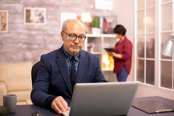 60 'lı yaşlarda yaşlı bir adam rahat oturma odasında dizüstü bilgisayarla çalışıyor. - Fotoğraf, Görsel
